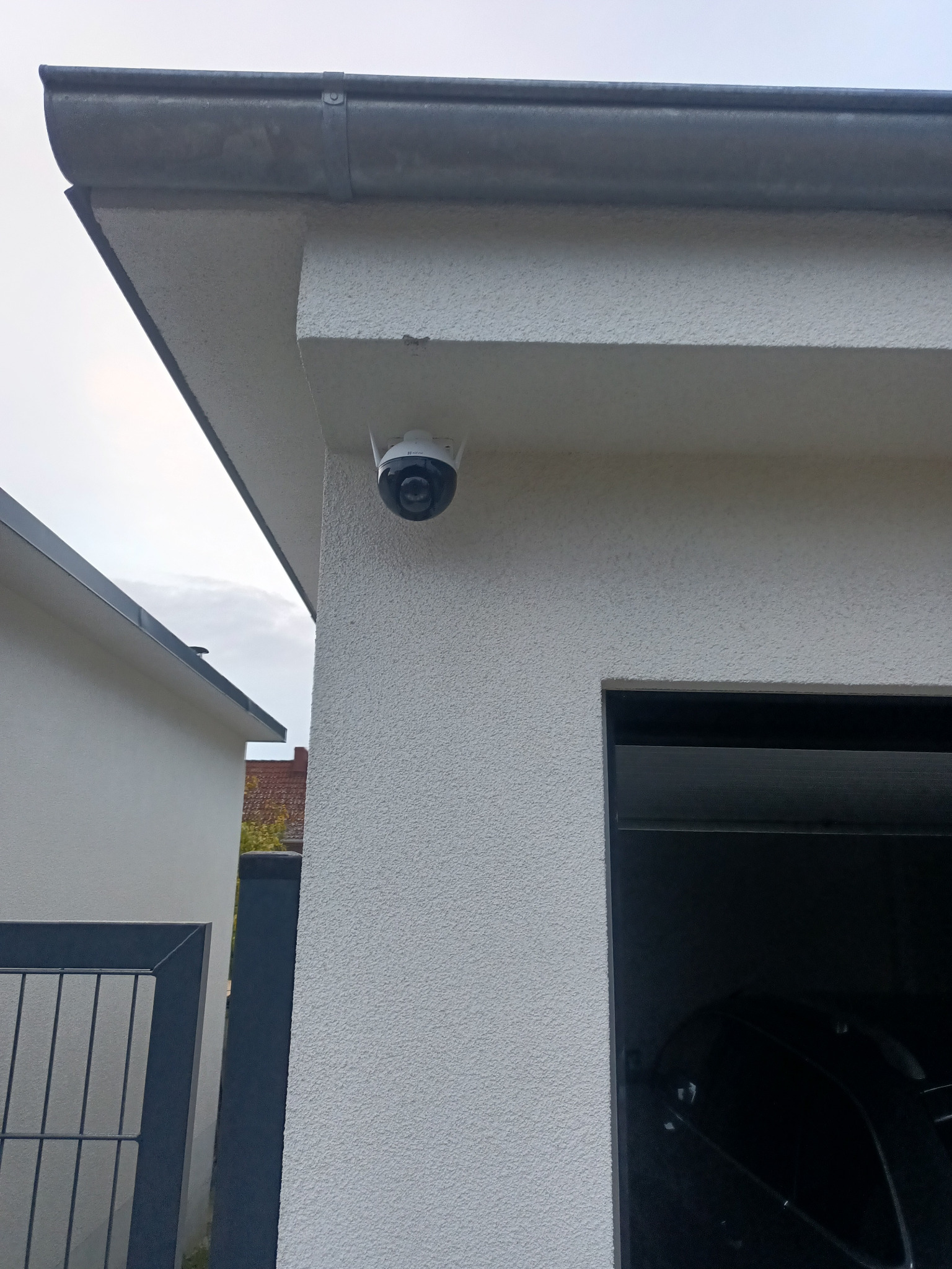 Kamera monitoringu zewnętrznego zamontowana na elewacji budynku w Szczecinie, widoczny fragment dachu i rynny, w tle brama wjazdowa i sąsiedni budynek.