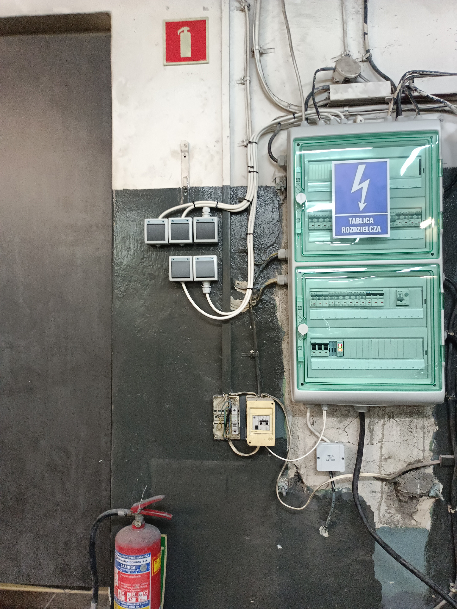 Dwu-segmentowa tablica rozdzielcza w obudowie z tworzywa sztucznego, zamontowana na pomalowanej ścianie z widocznymi śladami użytkowania, z podłączonymi przewodami. Obok widoczne trzy podwójne...