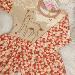 AW Designer Aleksandra Wisińska - Dziecięca sukienka z długim rękawem, beżowa góra z rysunkiem sarny i geometryczny wzór trójkątów na rękawach i spódnicy, leżąca na białym futrze.