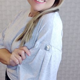 AW Designer Aleksandra Wisińska - Uśmiechnięta blondynka w okularach prezentuje szarą bluzę z plisowanym rękawem i metalowym logo na tle ściany z geometrycznym wzorem.