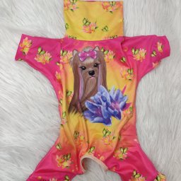 AW Designer Aleksandra Wisińska - Różowo-żółty kombinezon dla psa z nadrukiem psa w kokardzie, kwiatów i motyli, leżący na białym, puszystym tle.