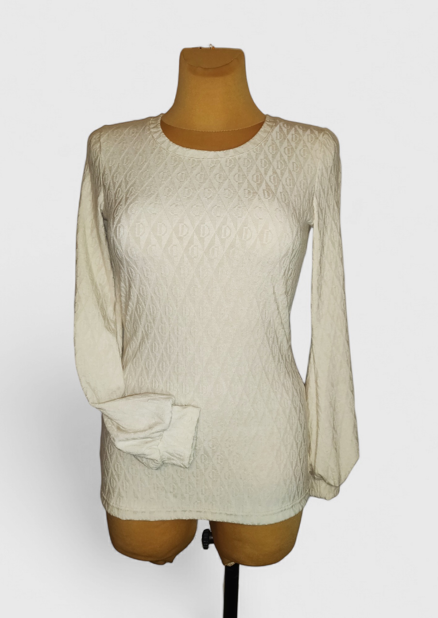 Kremowa bluzka z długim rękawem i wytłaczanym wzorem na manekinie krawieckim, widoczne detale tkaniny.