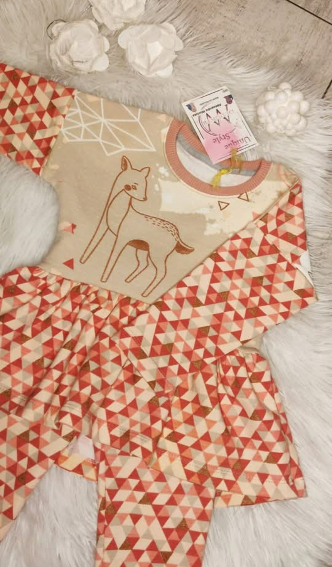 Dziecięca sukienka z długim rękawem, beżowa góra z rysunkiem sarny i geometryczny wzór trójkątów na rękawach i spódnicy, leżąca na białym futrze.