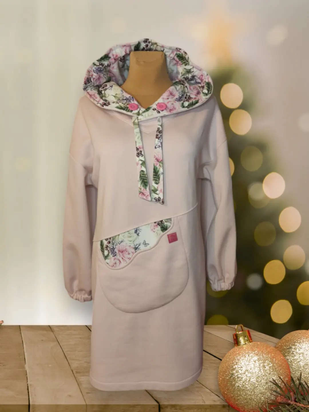 Bladoróżowa sukienka dresowa z kapturem i kieszenią, ozdobiona wzorem w kwiaty, prezentowana na manekinie na tle choinki z lampkami i złotymi bombkami.