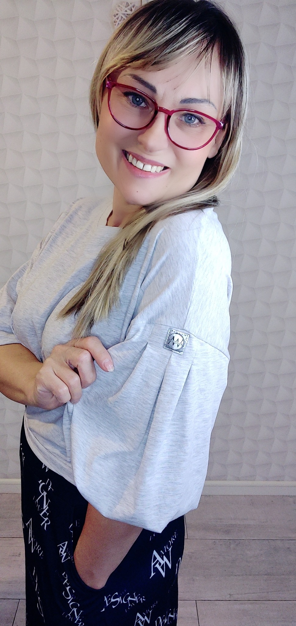 Uśmiechnięta blondynka w okularach prezentuje szarą bluzę z plisowanym rękawem i metalowym logo na tle ściany z geometrycznym wzorem.