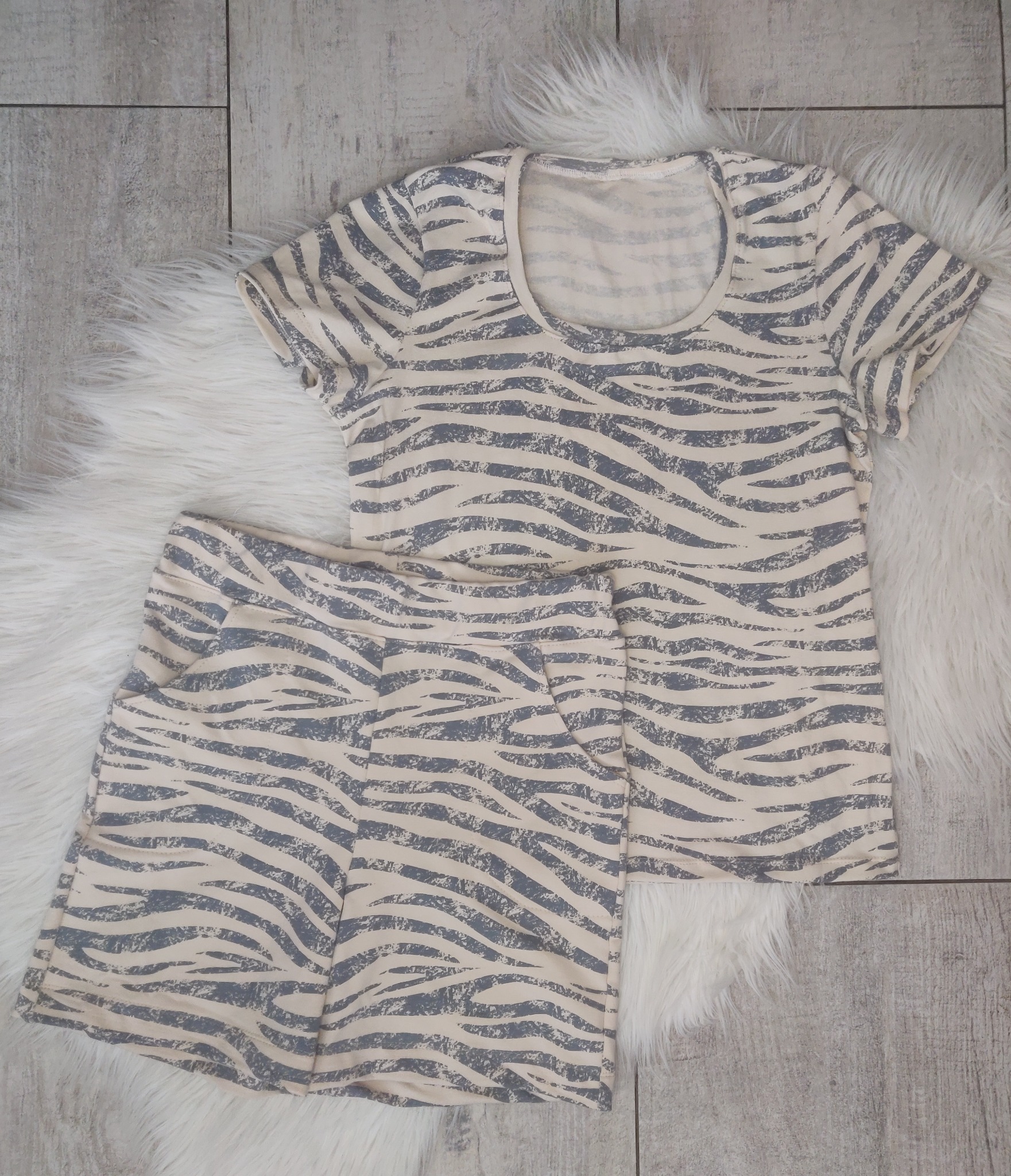 Komplet damski: beżowa koszulka z krótkim rękawem i szorty z wzorem zebry, leżące na białym, puszystym dywaniku na tle drewnopodobnych płytek.