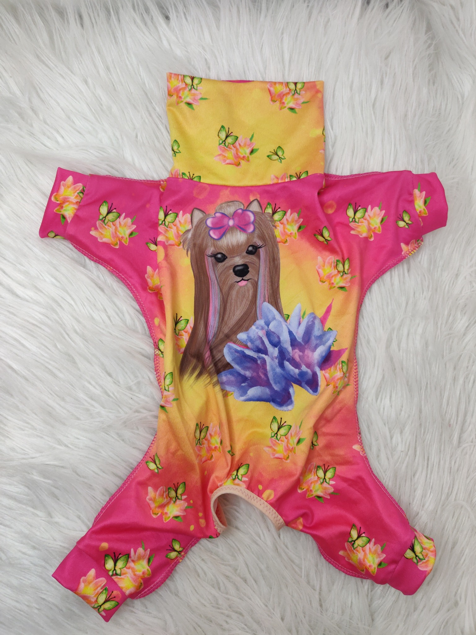 Różowo-żółty kombinezon dla psa z nadrukiem psa w kokardzie, kwiatów i motyli, leżący na białym, puszystym tle.