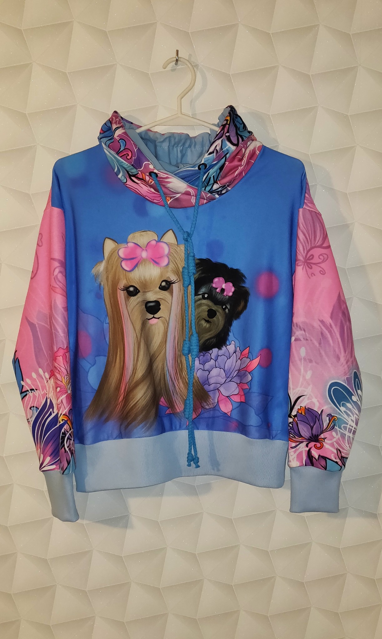 Niebiesko-różowa bluza z kapturem, z nadrukiem dwóch psów rasy Yorkshire Terrier z kokardkami, zawieszona na wieszaku na tle białej ściany z geometrycznym wzorem.