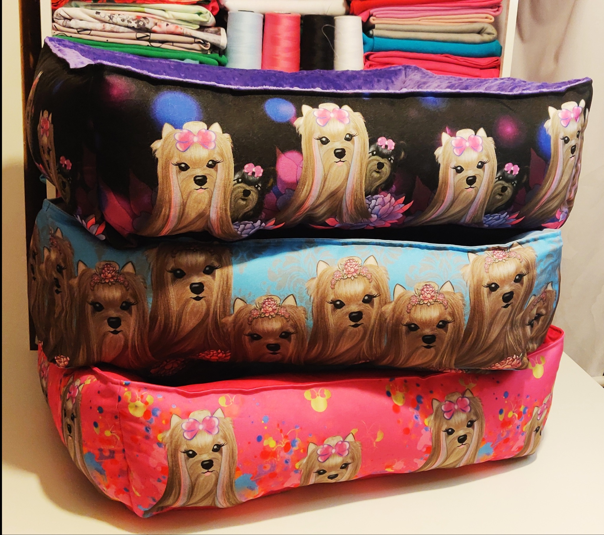 Stos trzech różnokolorowych, ręcznie szytych legowisk dla psów z nadrukiem wizerunków psów rasy yorkshire terrier w kokardkach, spiętrzone na białym stole, z materiałami i nićmi w tle.