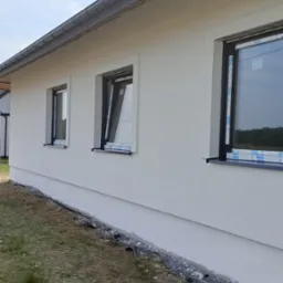 Świeżo otynkowana, biała elewacja domu jednorodzinnego z trzema czarnymi oknami z folią ochronną, widok z boku, częściowo widoczny system drenażowy przy fundamencie.
