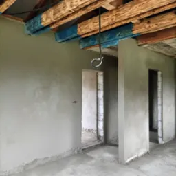 Surowe wnętrze w trakcie prac wykończeniowych, z świeżo nałożonym tynkiem na ścianach, drewnianymi belkami stropowymi częściowo pomalowanymi na niebiesko i widocznymi otworami drzwiowymi.