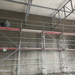 Szymon Jesiolkiewicz - Pracownicy na rusztowaniu podczas nakładania zaprawy na ścianę wewnątrz budynku z widoczną konstrukcją dachu