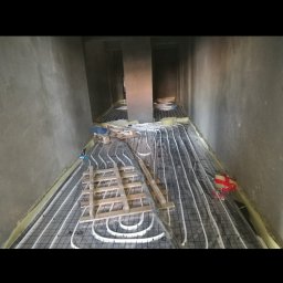 Szymon Jesiolkiewicz - Montaż ogrzewania podłogowego w surowym wnętrzu, widoczne rury ułożone na siatce, drewniane podpory i ściany w stanie deweloperskim.