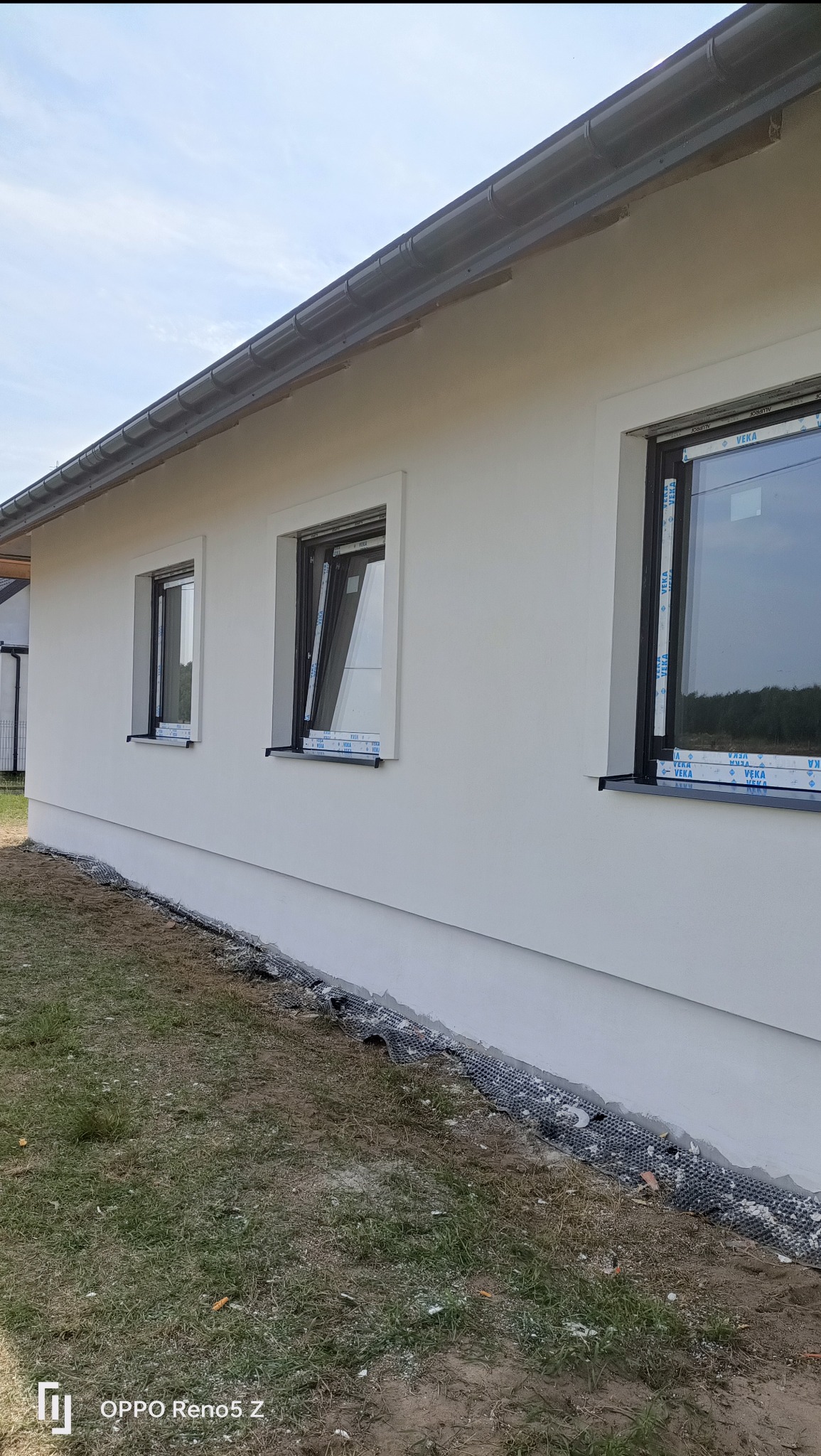 Świeżo otynkowana, biała elewacja domu jednorodzinnego z trzema czarnymi oknami z folią ochronną, widok z boku, częściowo widoczny system drenażowy przy fundamencie.