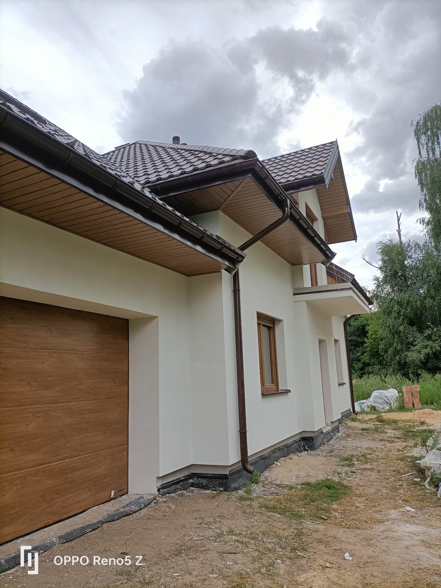 Nowoczesny dom jednorodzinny z jasną elewacją, brązową stolarką okienną i bramą garażową, widok z boku na tle pochmurnego nieba i zieleni drzew.