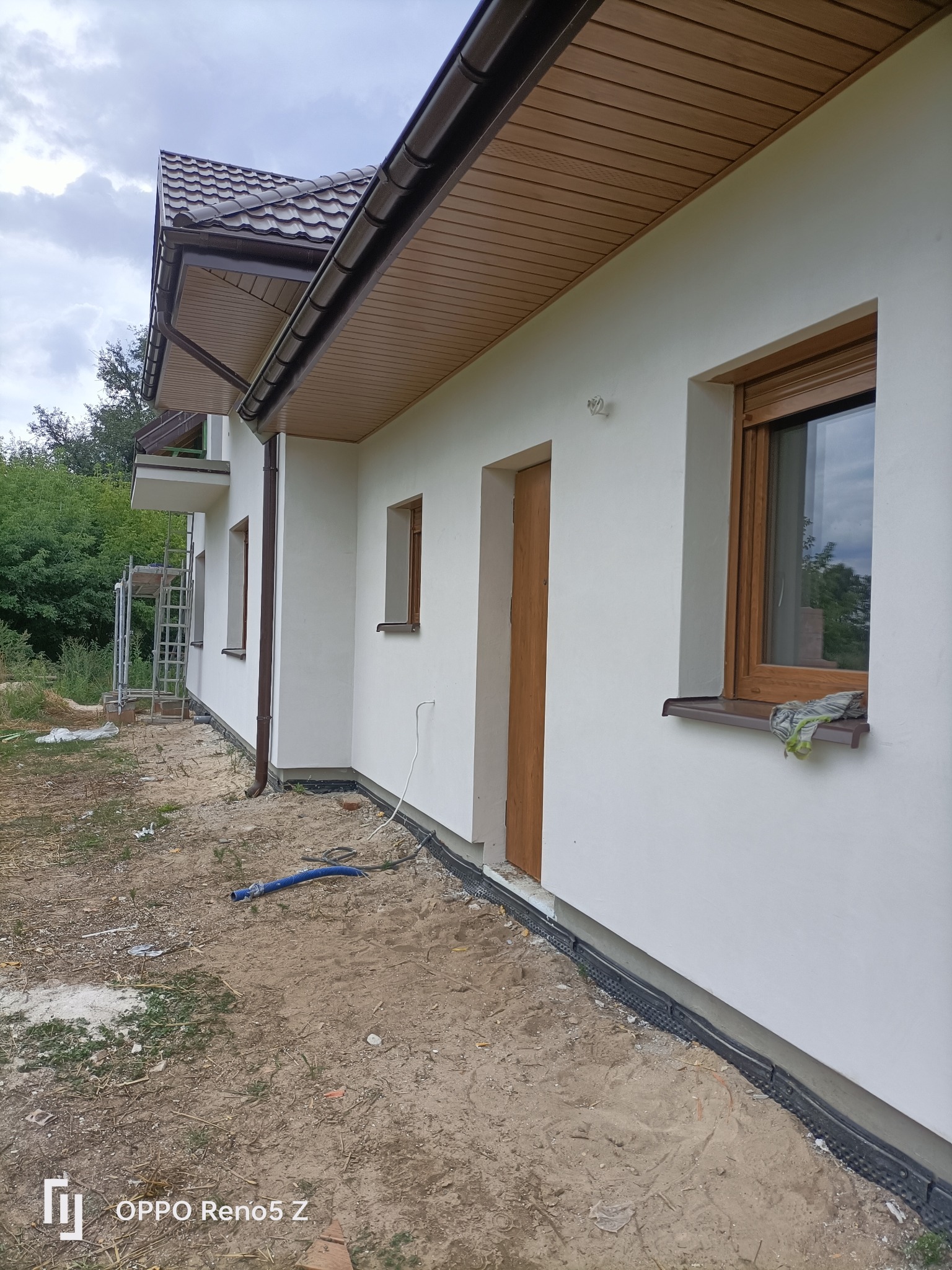 Świeżo otynkowana elewacja domu jednorodzinnego w kolorze białym, z widocznymi drewnianymi oknami i drzwiami, na tle pochmurnego nieba, z elementami rusztowania w tle.