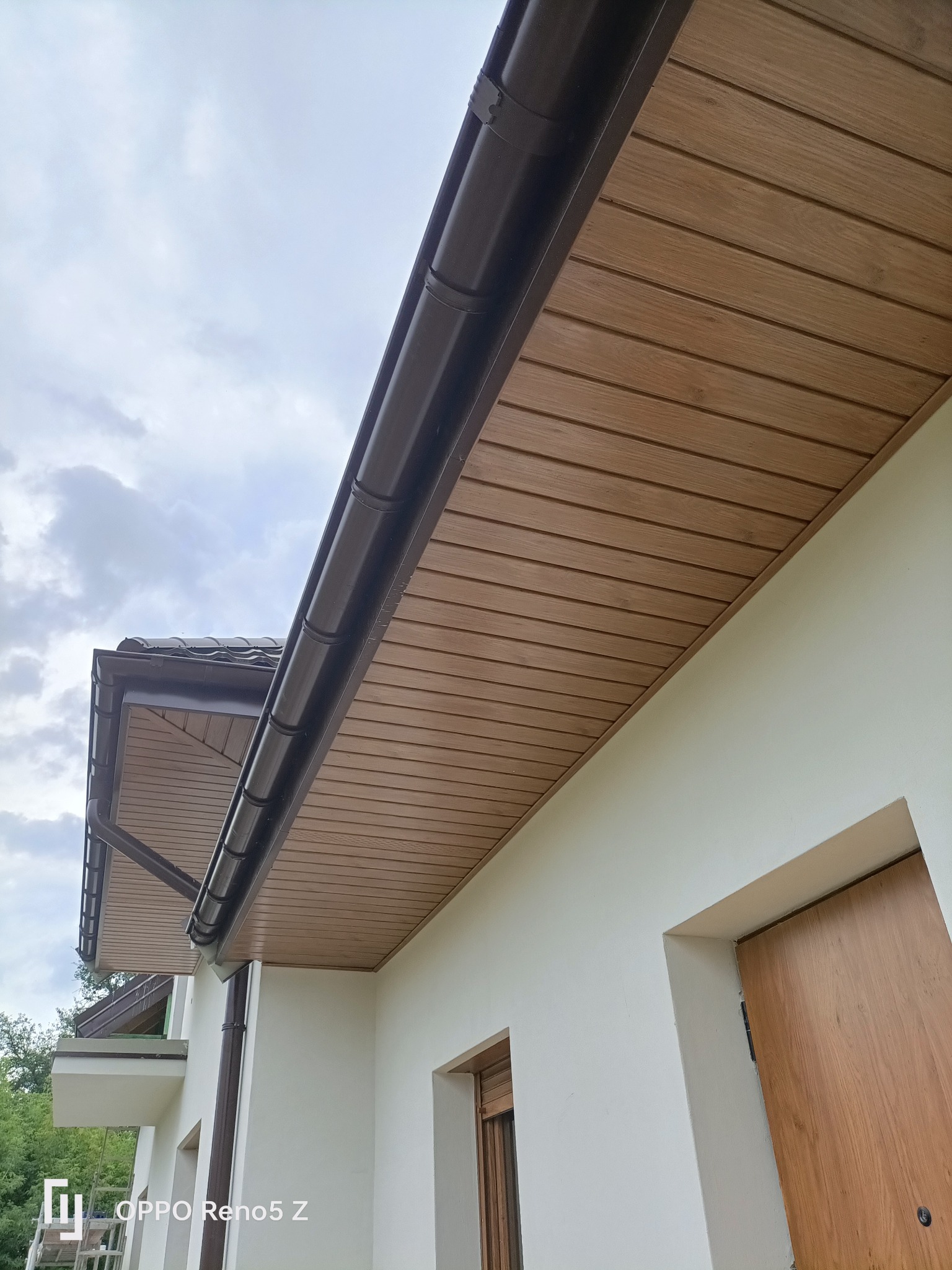 Drewnopodobna podbitka dachowa z brązowymi rynnami zamontowana na elewacji domu, widok z dołu w pochmurny dzień.