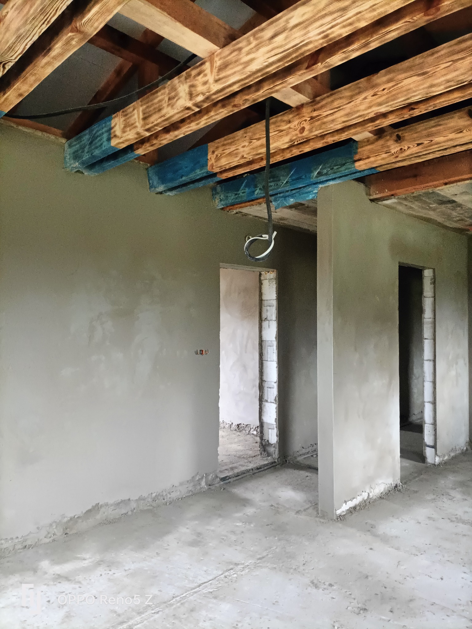 Surowe wnętrze w trakcie prac wykończeniowych, z świeżo nałożonym tynkiem na ścianach, drewnianymi belkami stropowymi częściowo pomalowanymi na niebiesko i widocznymi otworami drzwiowymi.