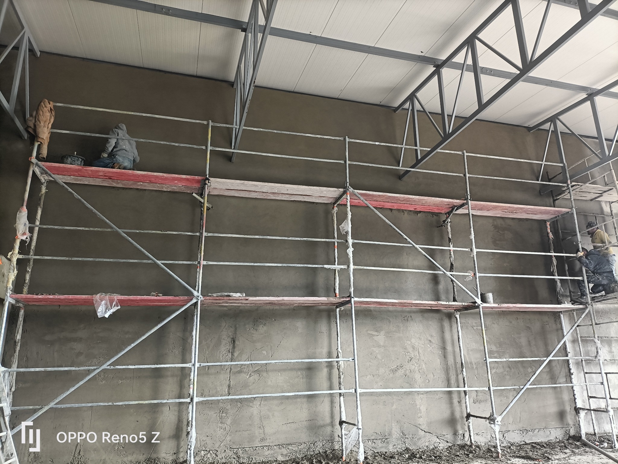 Pracownicy na rusztowaniu podczas nakładania zaprawy na ścianę wewnątrz budynku z widoczną konstrukcją dachu