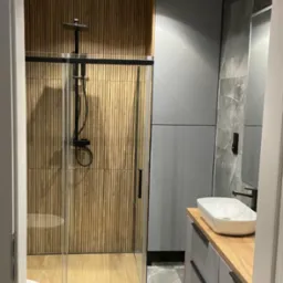 Nowoczesna łazienka z kabiną prysznicową typu walk-in z czarną armaturą, drewnianą ścianą z paneli pionowych, szarą szafką z umywalką nablatową i marmurową posadzką.