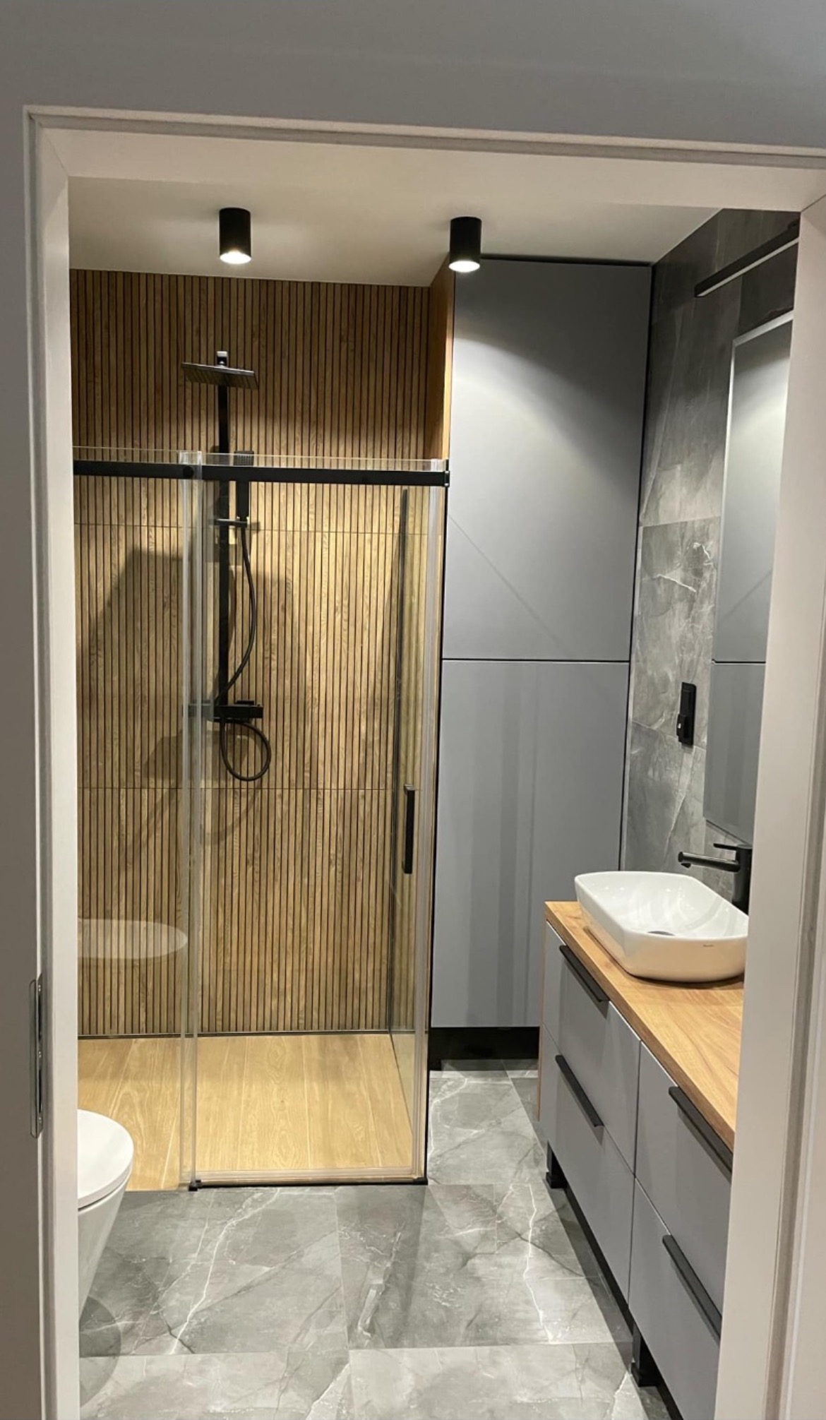 Nowoczesna łazienka z kabiną prysznicową typu walk-in z czarną armaturą, drewnianą ścianą z paneli pionowych, szarą szafką z umywalką nablatową i marmurową posadzką.