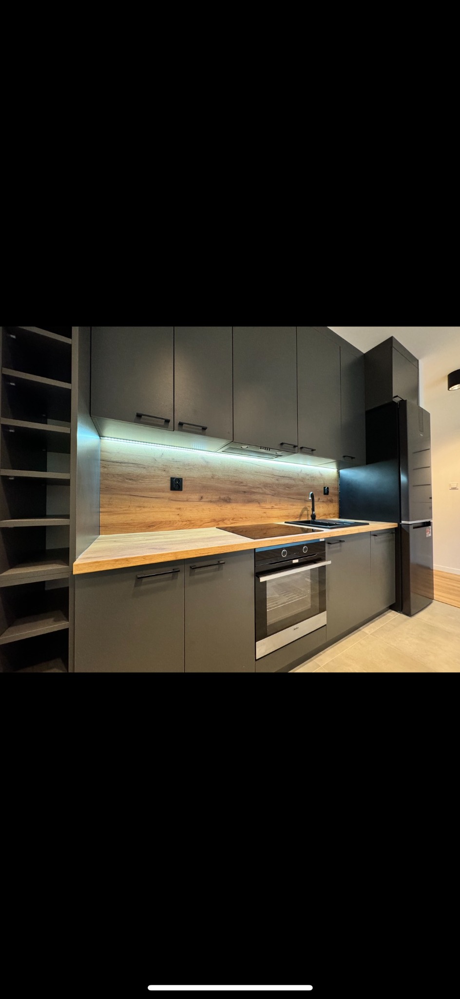 Nowoczesna kuchnia z szarymi szafkami, drewnianym blatem, podświetleniem LED i czarnym sprzętem AGD, w tym płyta indukcyjna i piekarnik.