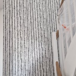 P.S. Majster Expert - Ściana pokryta tapetą z motywem brzozowego lasu, widoczne fragmenty białych schodów z zabezpieczonymi stopniami, w trakcie prac wykończeniowych.