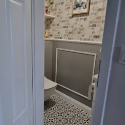 Wykończona łazienka w stylu angielskim z toaletą, wzorzystymi płytkami podłogowymi, ściana z efektem cegły i plakatem z Big Benem, widok zza uchylonych drzwi.