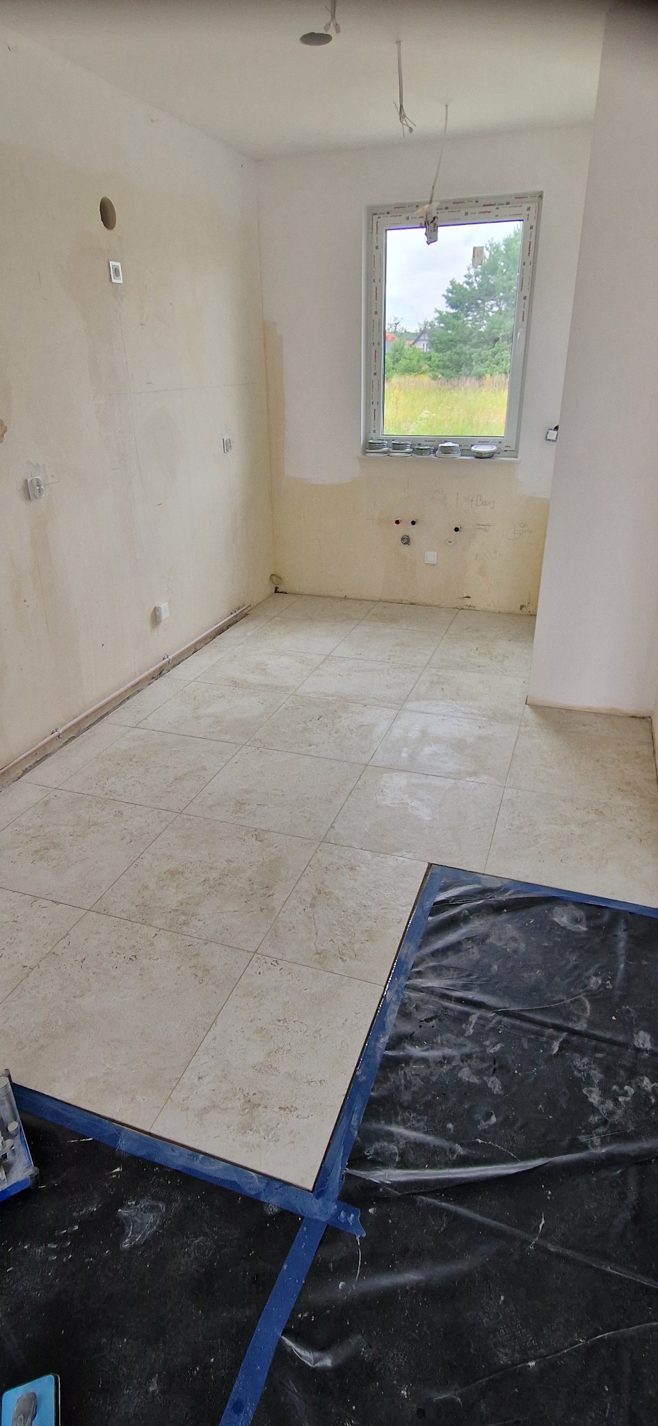 Układanie beżowych płytek podłogowych w pomieszczeniu w Łochowie, zabezpieczonym folią i taśmą malarską, ściany przygotowane do dalszych prac wykończeniowych, widoczne instalacje.