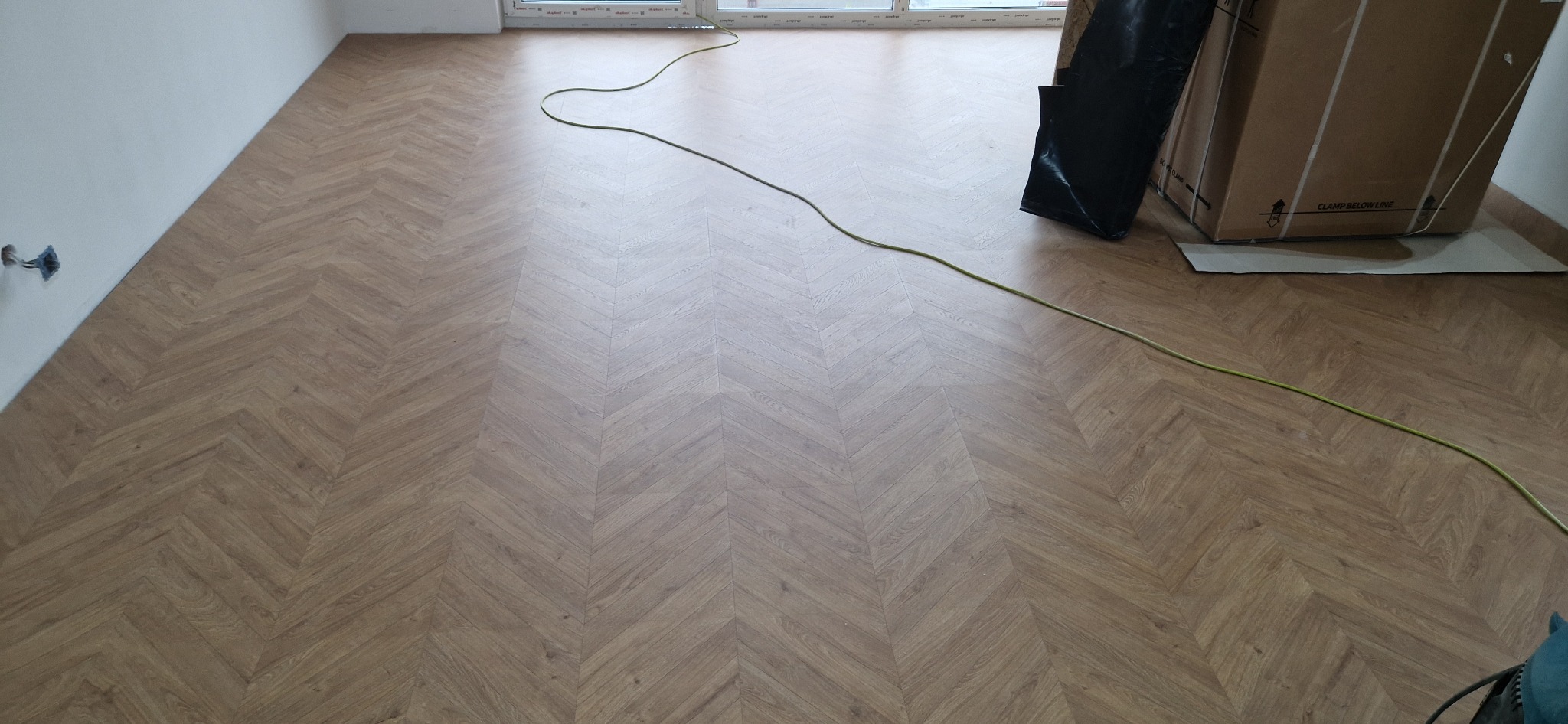 Ułożone panele podłogowe w jodełkę francuską, widoczny kabel zasilający narzędzie, kartonowe opakowanie paneli i fragment ściany z puszką elektryczną.