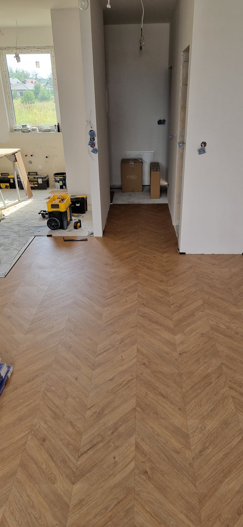 Ułożone panele podłogowe w jodełkę w trakcie remontu, widoczne narzędzia i puszki z farbą w tle, surowe ściany i instalacje elektryczne.