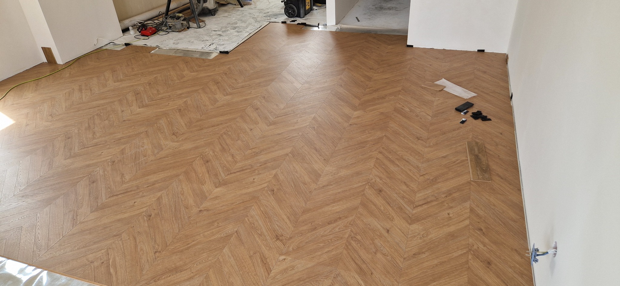 Układanie paneli podłogowych w jodełkę, widoczne narzędzia i elementy montażowe, fragment folii ochronnej na podłodze, pomieszczenie w trakcie prac wykończeniowych.