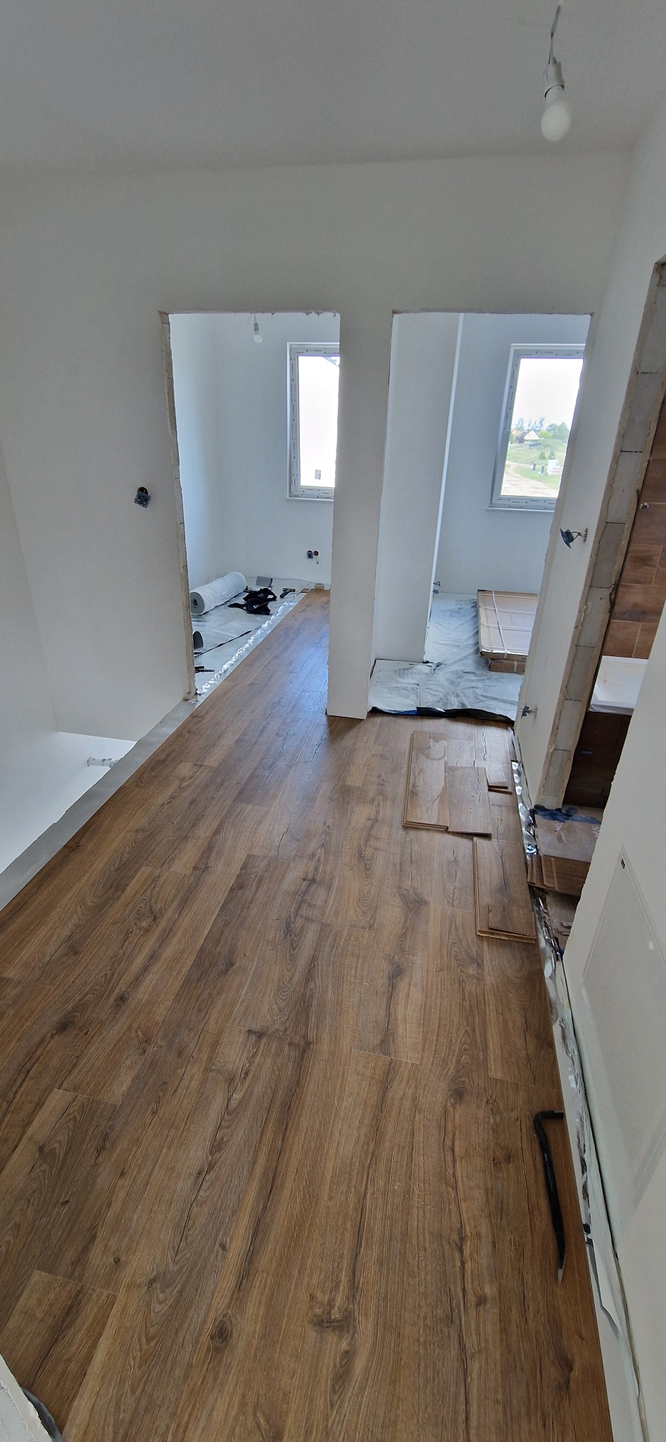 Układanie paneli podłogowych w jasnym wnętrzu, widoczne podkłady i paczki paneli w sąsiednich pomieszczeniach, podważka przy ścianie.