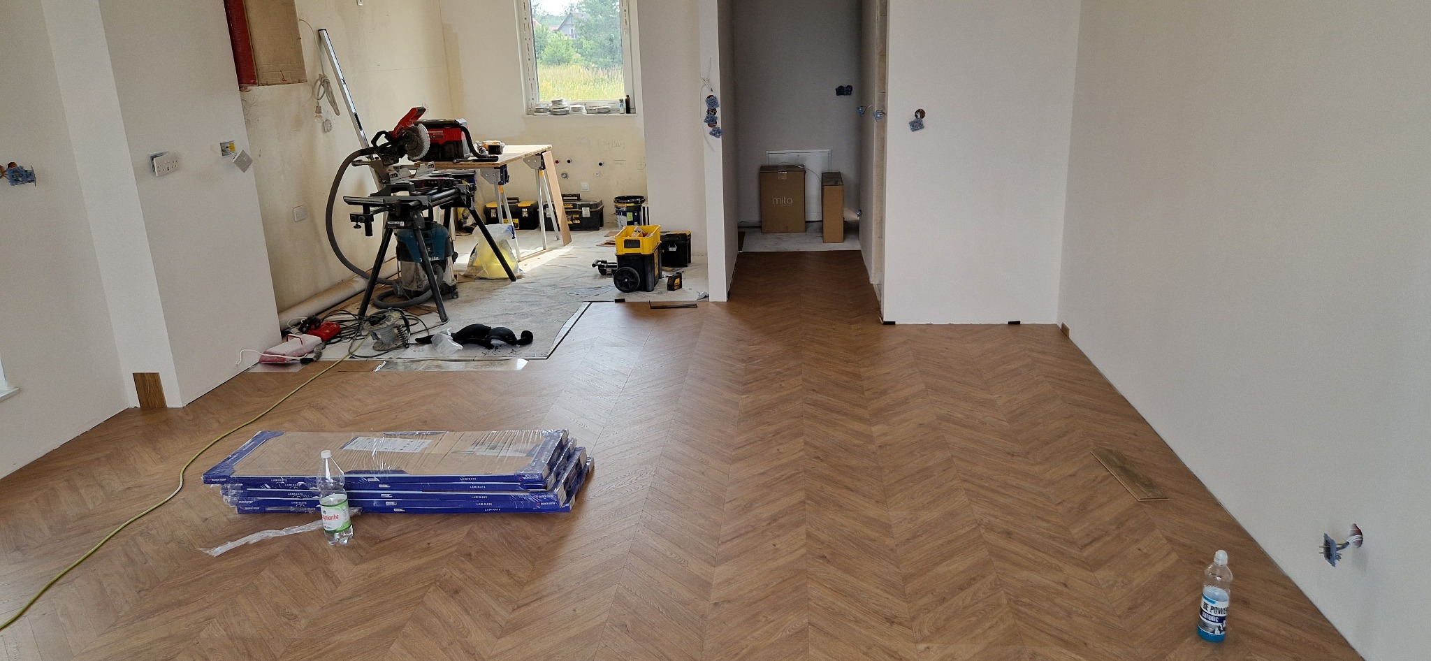 Układanie paneli w jodełkę w trakcie prac wykończeniowych, widoczne paczki paneli, narzędzia i butelka wody mineralnej.