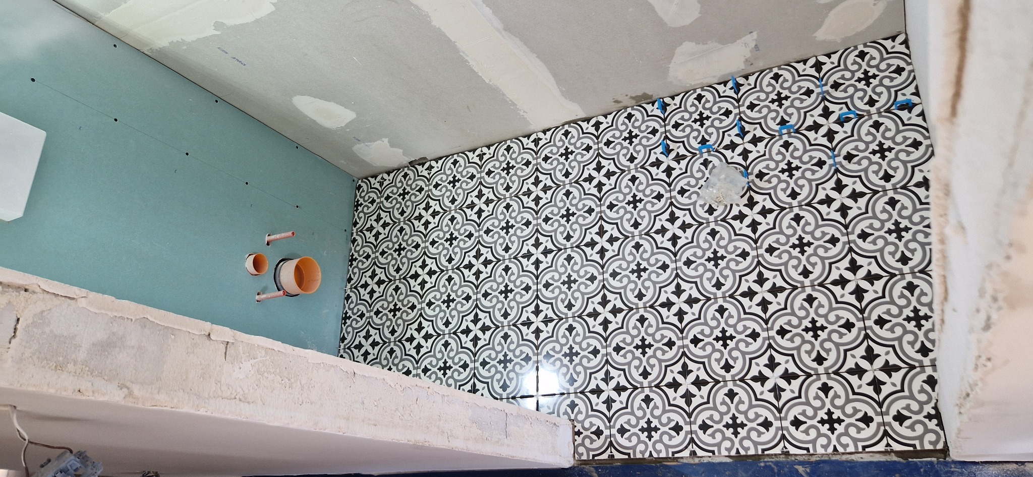 Ściana łazienki w trakcie wykańczania z ułożonymi wzorzystymi płytkami w odcieniach szarości i czerni, widoczne piony instalacyjne oraz płyta kartonowo-gipsowa na sąsiedniej ścianie...