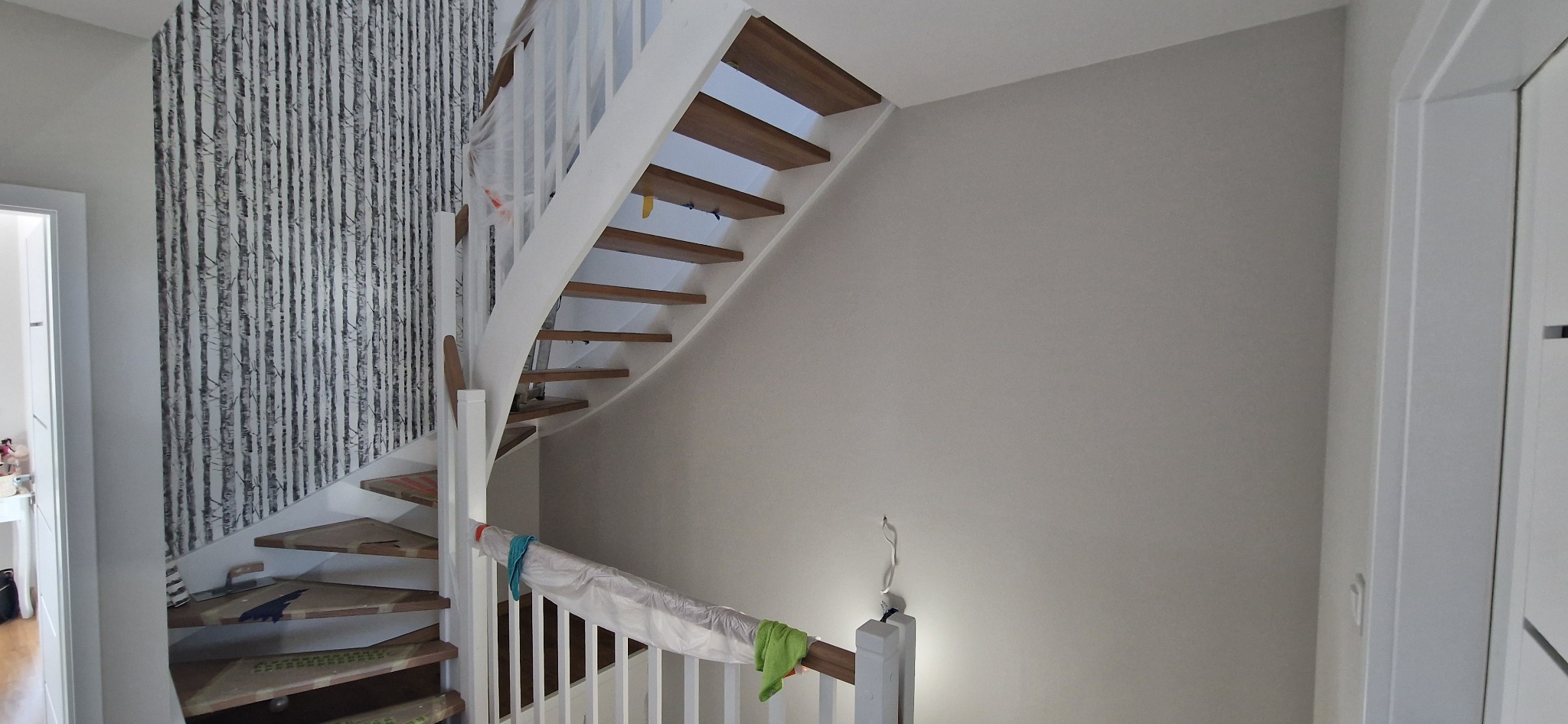 Wnętrze domu w trakcie prac wykończeniowych: drewniane schody z białymi balustradami, zabezpieczone folią i taśmą malarską, ściana z tapetą imitującą brzozowy las oraz świeżo pomalowana szara...