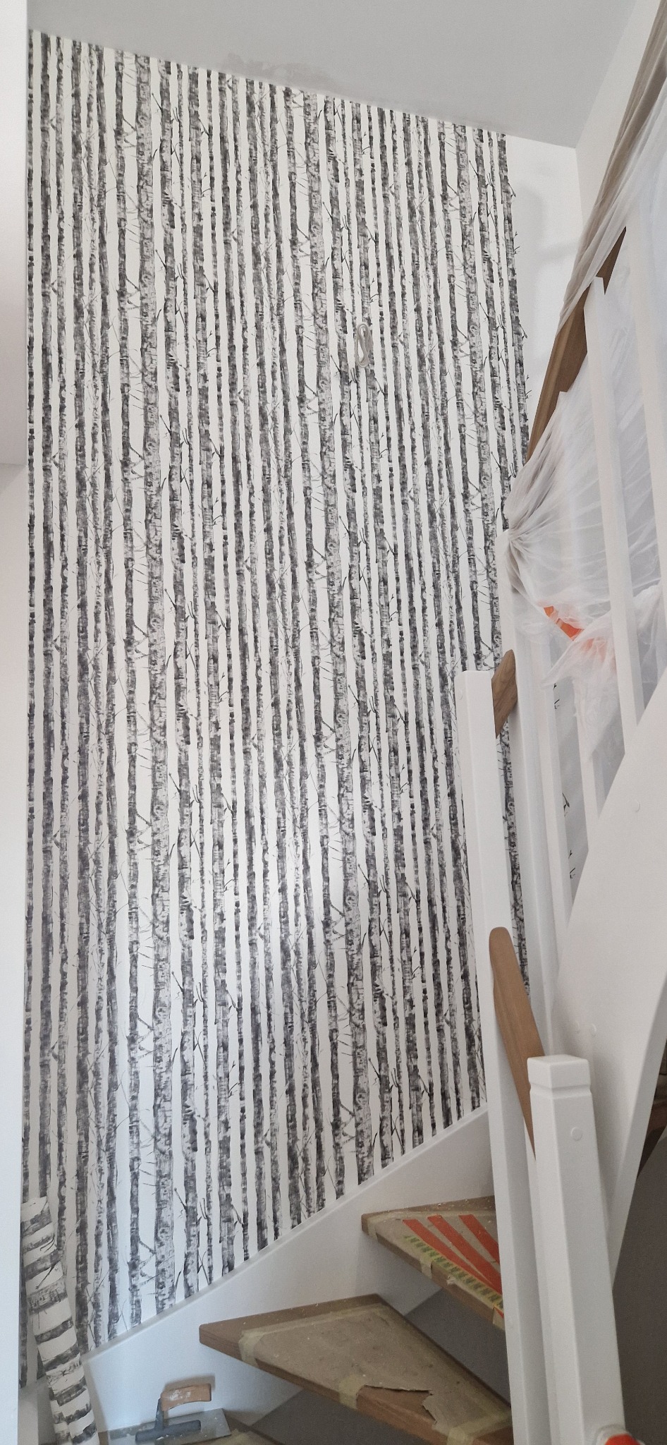 Ściana pokryta tapetą z motywem brzozowego lasu, widoczne fragmenty białych schodów z zabezpieczonymi stopniami, w trakcie prac wykończeniowych.