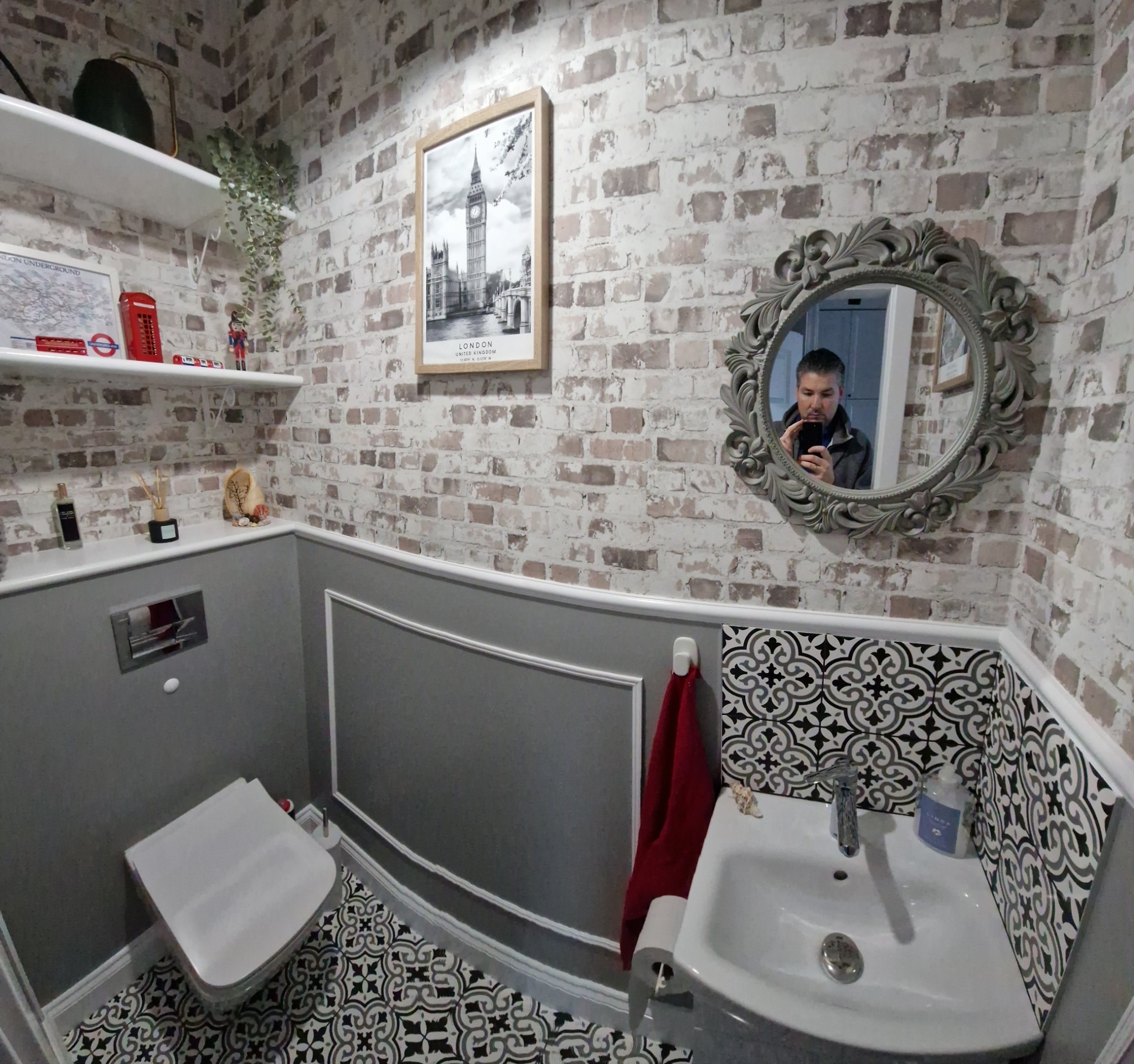 Wykończona łazienka z toaletą i umywalką, ściany wyłożone cegłą dekoracyjną i czarno-białymi płytkami wzorzystymi, lustro w ozdobnej ramie, widoczne akcenty angielskie: zdjęcie Big Bena w ramce...