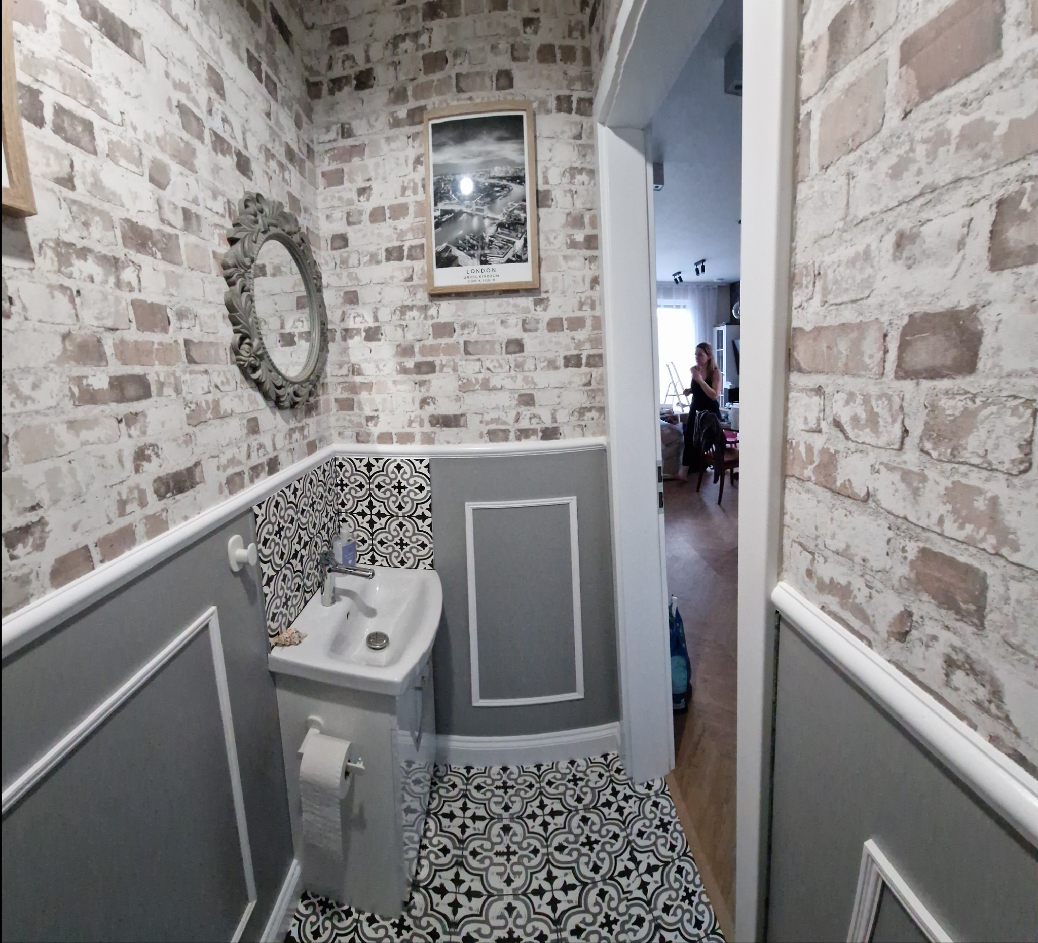 Mała łazienka w stylu angielskim z umywalką na postumencie, toaletą i dekoracyjnymi płytkami podłogowymi w czarno-białe wzory. Ściany wykończone cegłą i szarą boazerią.