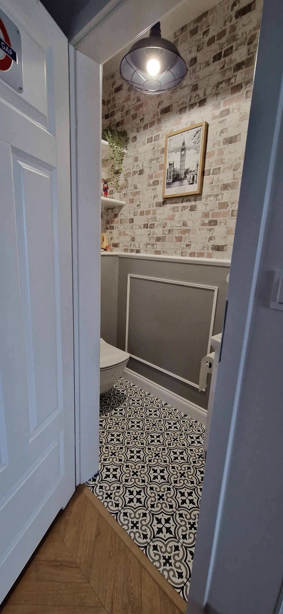 Wykończona łazienka w stylu angielskim z toaletą, wzorzystymi płytkami podłogowymi, ściana z efektem cegły i plakatem z Big Benem, widok zza uchylonych drzwi.