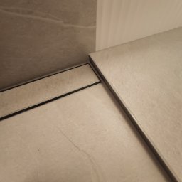 Andrzej Jeleń - Szczegółowe ujęcie narożnika wykończonego beżowymi płytkami z widocznym odpływem liniowym, przy białej ścianie.