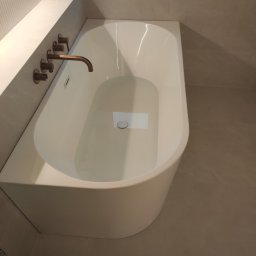 Andrzej Jeleń - Nowoczesna, biała wanna wolnostojąca wypełniona wodą, z miedzianą armaturą w ścianie, na tle płytek w odcieniach beżu i szarości.