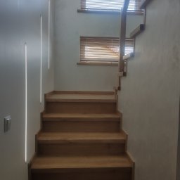 Andrzej Jeleń - Drewniane schody z minimalistyczną balustradą, oświetlenie LED pionowe w ścianie, widoczne żaluzje drewniane w oknach.