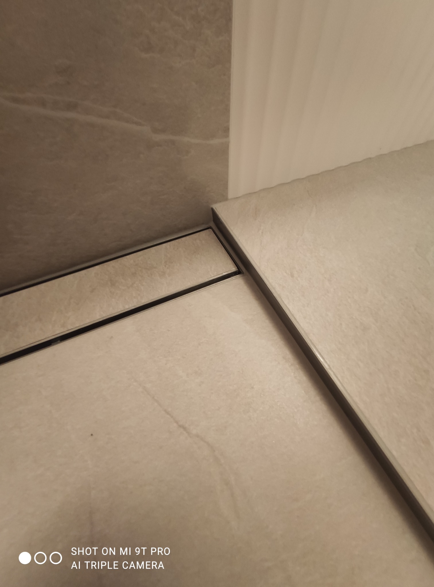 Szczegółowe ujęcie narożnika wykończonego beżowymi płytkami z widocznym odpływem liniowym, przy białej ścianie.