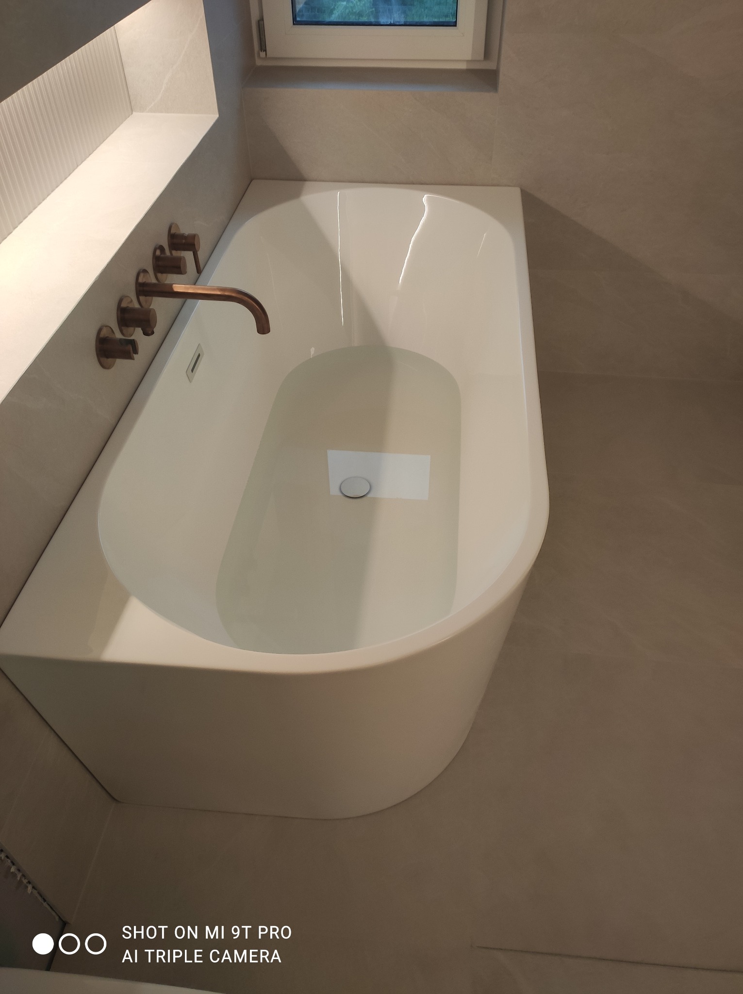 Nowoczesna, biała wanna wolnostojąca wypełniona wodą, z miedzianą armaturą w ścianie, na tle płytek w odcieniach beżu i szarości.