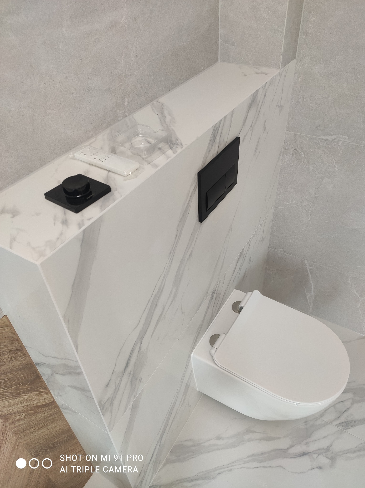 Elegancka toaleta podwieszana w Borkach, wykończona płytkami imitującymi marmur, z czarnym przyciskiem spłukującym i półką na akcesoria.