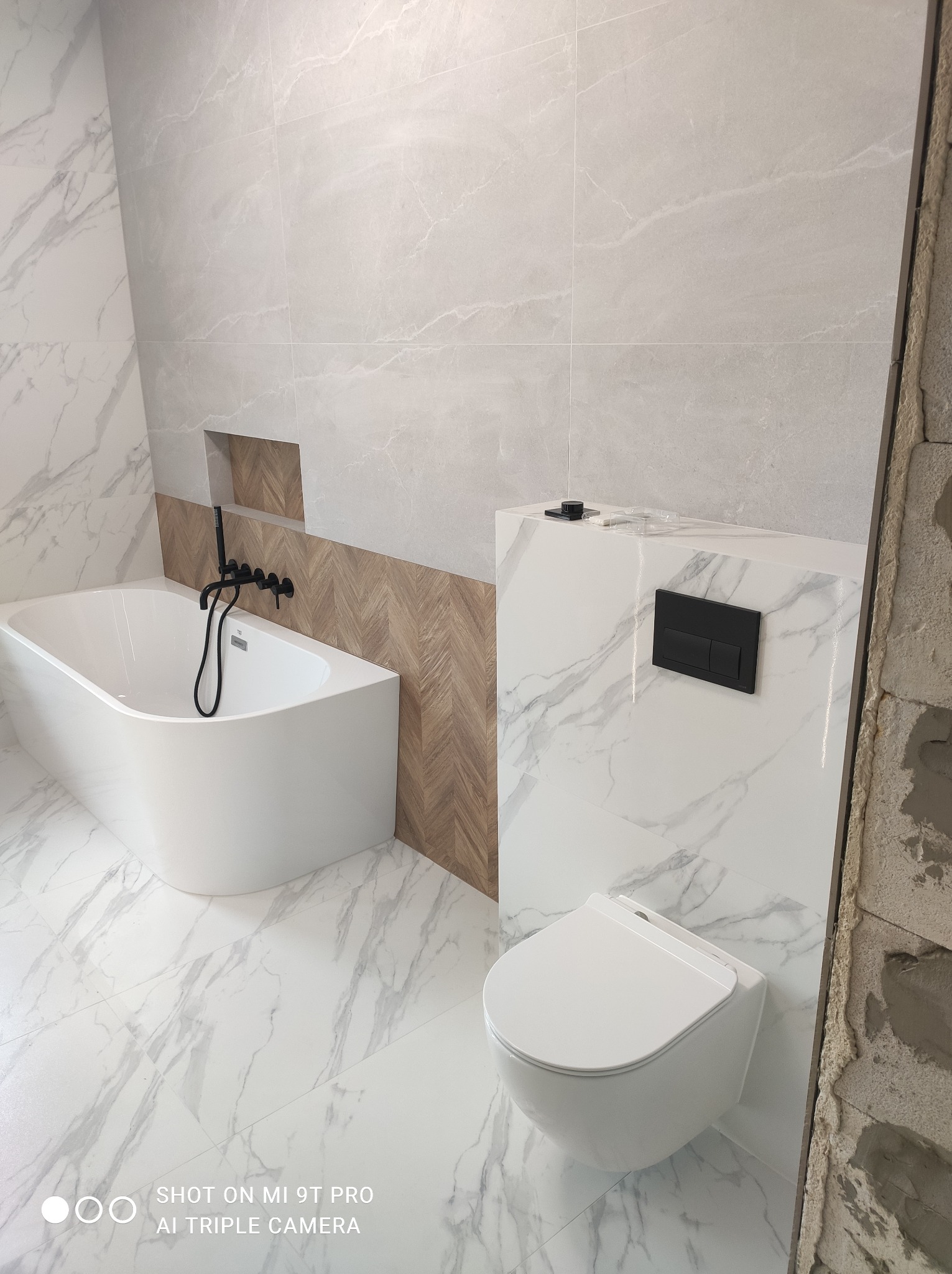 Nowoczesna łazienka w trakcie wykańczania: biała wanna z czarną armaturą, podwieszana toaleta, marmurowe płytki na podłodze i ścianach, drewnopodobna mozaika w niszy.