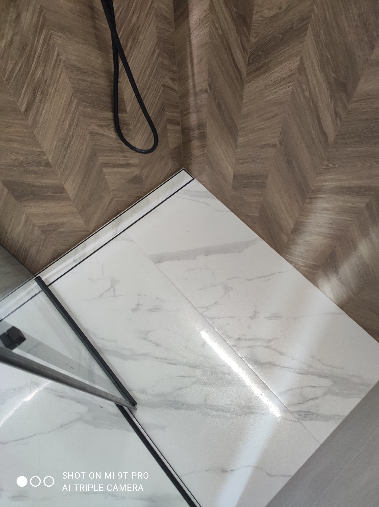 Połączenie płytek podłogowych imitujących drewno w jodełkę z białymi płytkami imitującymi marmur, oddzielone czarną fugą, widoczne elementy kabiny prysznicowej z czarnymi profilami.