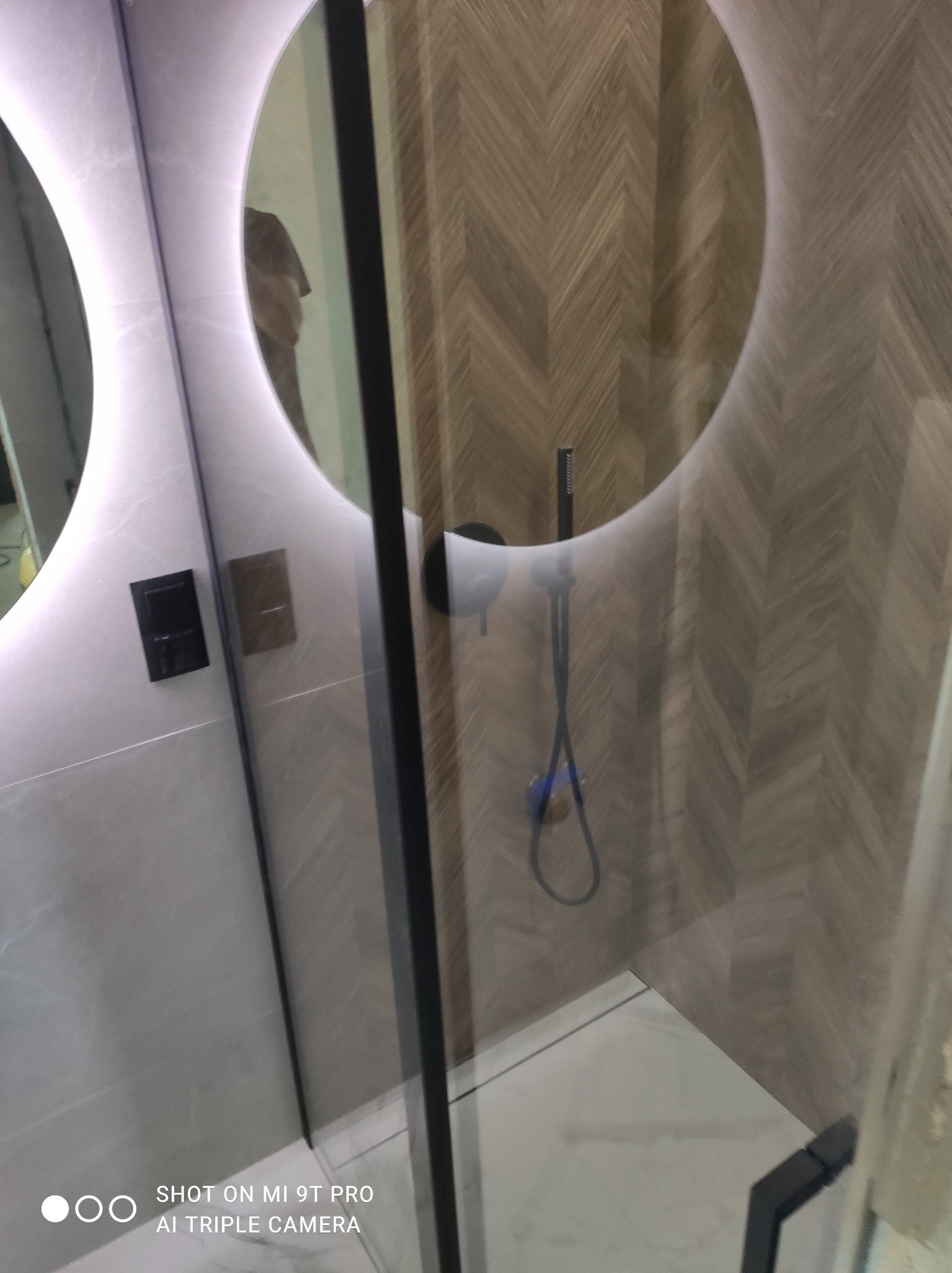 Nowoczesna łazienka z okrągłym lustrem podświetlanym LED, kabiną prysznicową typu walk-in i ściany wykończone płytkami imitującymi drewno ułożonymi w jodełkę.