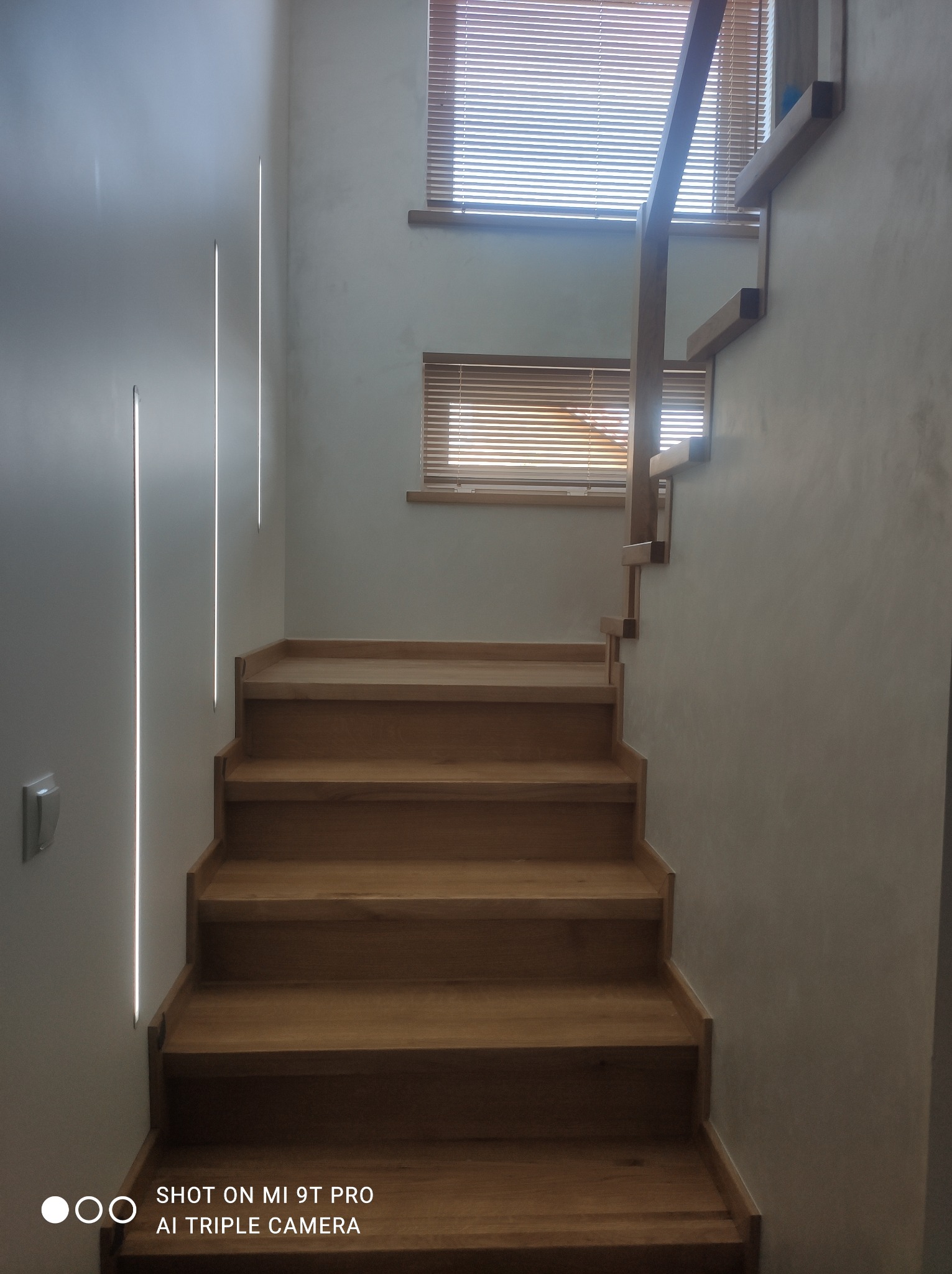 Drewniane schody z minimalistyczną balustradą, oświetlenie LED pionowe w ścianie, widoczne żaluzje drewniane w oknach.